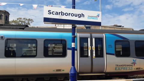 A blue TransPennine Express train at the platform in Scarborough. A sign reading Scarborough is attached to a pole, bunting can be seen in the background.