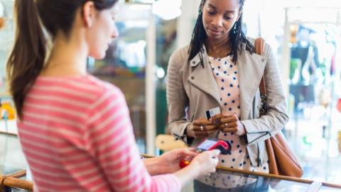 A woman in a shop holds her bank card ready to pay for an item while a shop worker holds the card machine