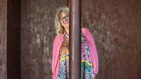 Anya Gallaccio at her installation in Paisley - she is standing in a room where the walls are painted with chocolate and she is wearing a floral dress and pink cardigan