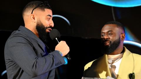 James Duncan and Fuhad Dawodu receiving a Mobo Award in February. James has short hair and a black beard, he wears sunglasses on his head and a blue suit, holding a mic. He's looking at Fuhad, who also has short hair and a black beard and is wearing a greeny-yellow suit over a white top. The staging behind them is black with streaks of blue.  