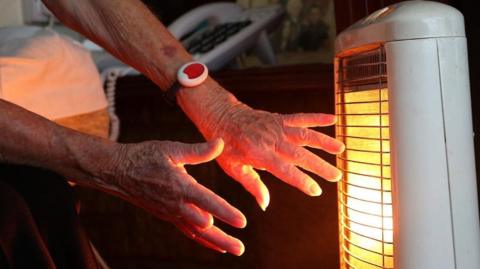 An elderly woman warms her hands at an electric fire