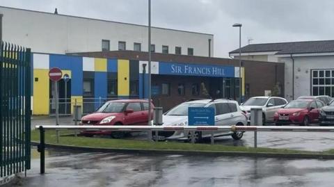 An image of the front of the school, with cars parked up outside. It has colourful plastic cladding.