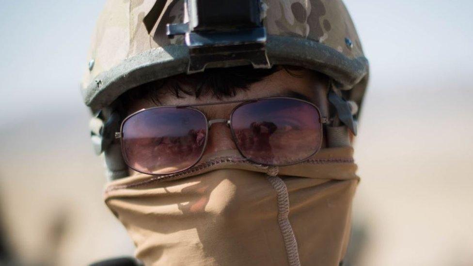 A members of the Triples pictured front on with his head in shot. He is wearing a combat helmet, sunglasses, and his mouth is covered