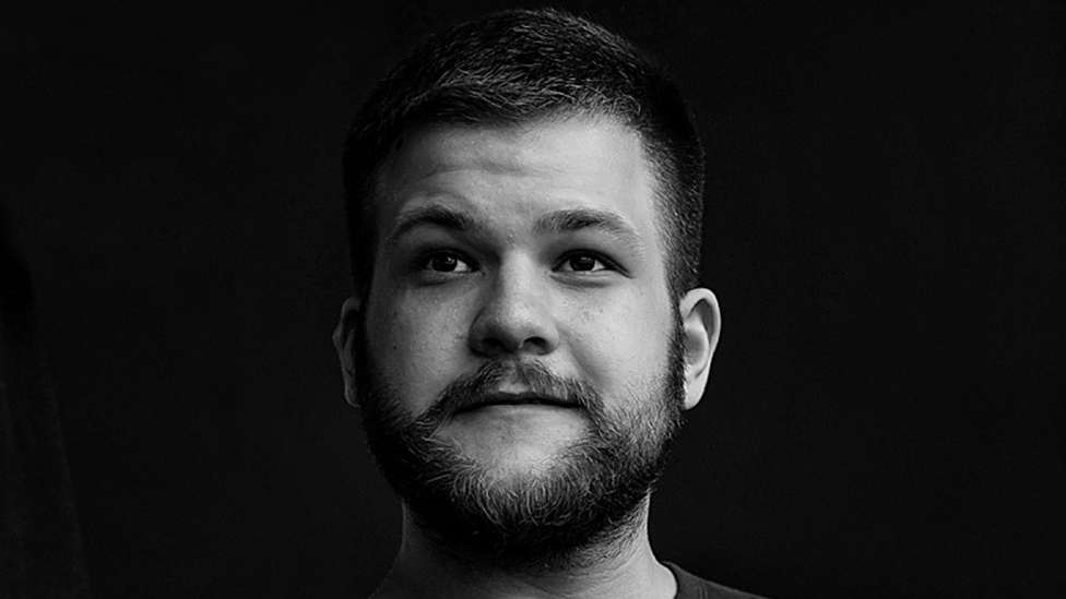 A black-and-white image of a man with black hair and beard looking into the middle distance. He's got a black background behind him and is artistically lit from the side, his face gradually falling into shadow from right to left.