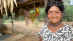 Tsimane woman with dark hair pinned up, wearing a white top with black flowers and a necklace