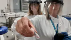 One woman wear white lab coat, hold small capsule wey contain blood. One woman stand behind her shoulder. Both wear PPE face guards.