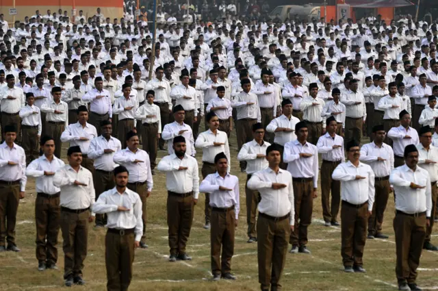 Des rangées d'hommes se tiennent en ligne organisée, le bras droit en travers de la poitrine et la paume de la main à plat, ce qui constitue la signature du salut RSS.