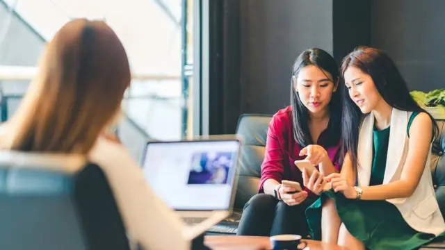 Two young women, sitting on a sofa close to each other and looking at a smartphone's screen together. Opposite them, there's a lone woman with laptop. It looks like they are not close friends, despite sharing a table at a cafe.