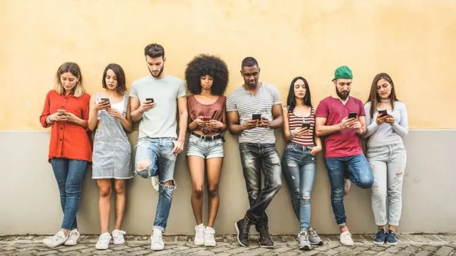 A group of young adults, all leaning against an ochre coloured wall, texting on their phones