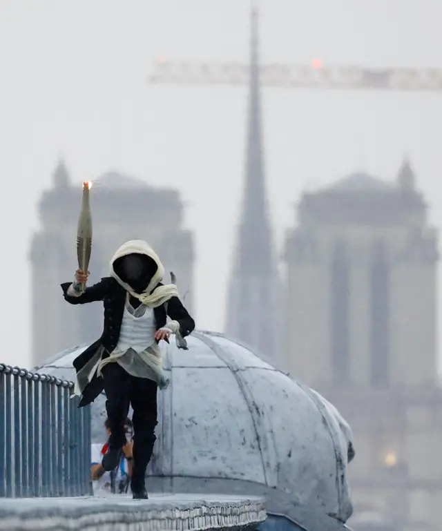  A  masked torchbearer runs atop the Musee d'Orsay with the Notre-Dame-de-Paris cathedral in the background during the Opening Ceremony of the Olympic Games Paris 2024 on July 26, 2024