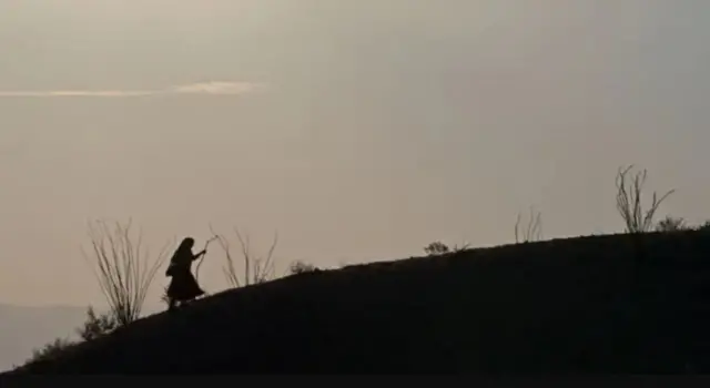 Image de la silhouette d'une femme portant une jupe et munie d'un bâton, marchant sur une pente.