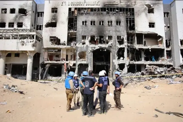 Des hommes debout devant la façade détruite  de l’hôpital Al Shifa