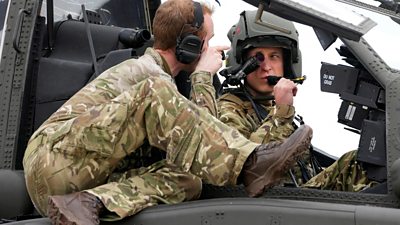 Prince William wearing army fatigues and a helmet sits in an Apache helicopter