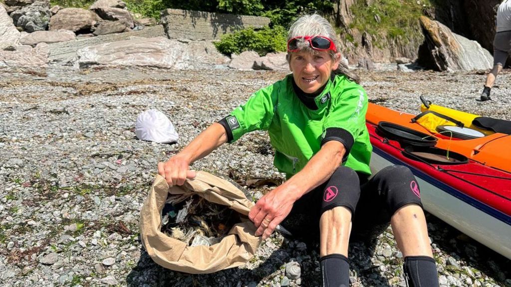 Kayaker Phillipa Arding, known as Pa, sits on a stony beach with a large paper bag full of rubbish she has collected