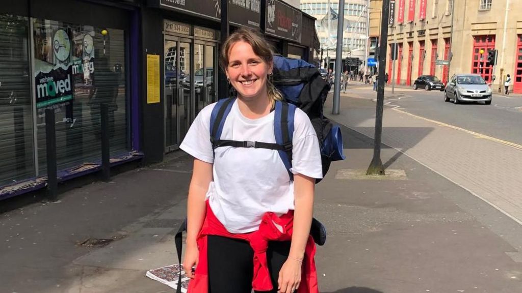 Chloe Davies has her hair tied back and is wearing a white t-shirt and black bottoms, with a red jacket tied around her waist. She has a large blue and black backpack on her back. She is smiling at the camera as she stands on the pavement next to an urban road