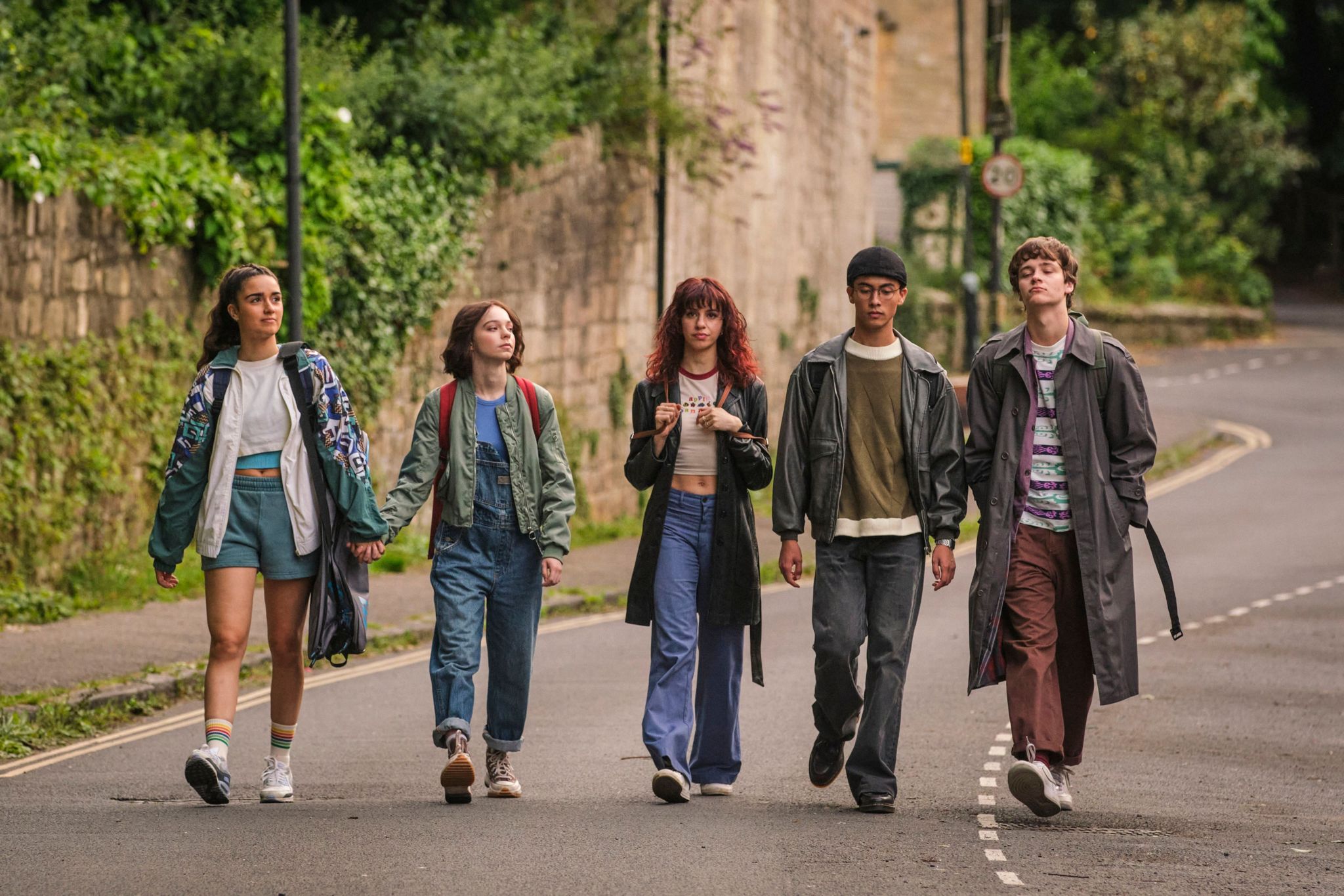 A group of five young people, three women and two men, are walking in line on the street, with two of the girls are holding hands