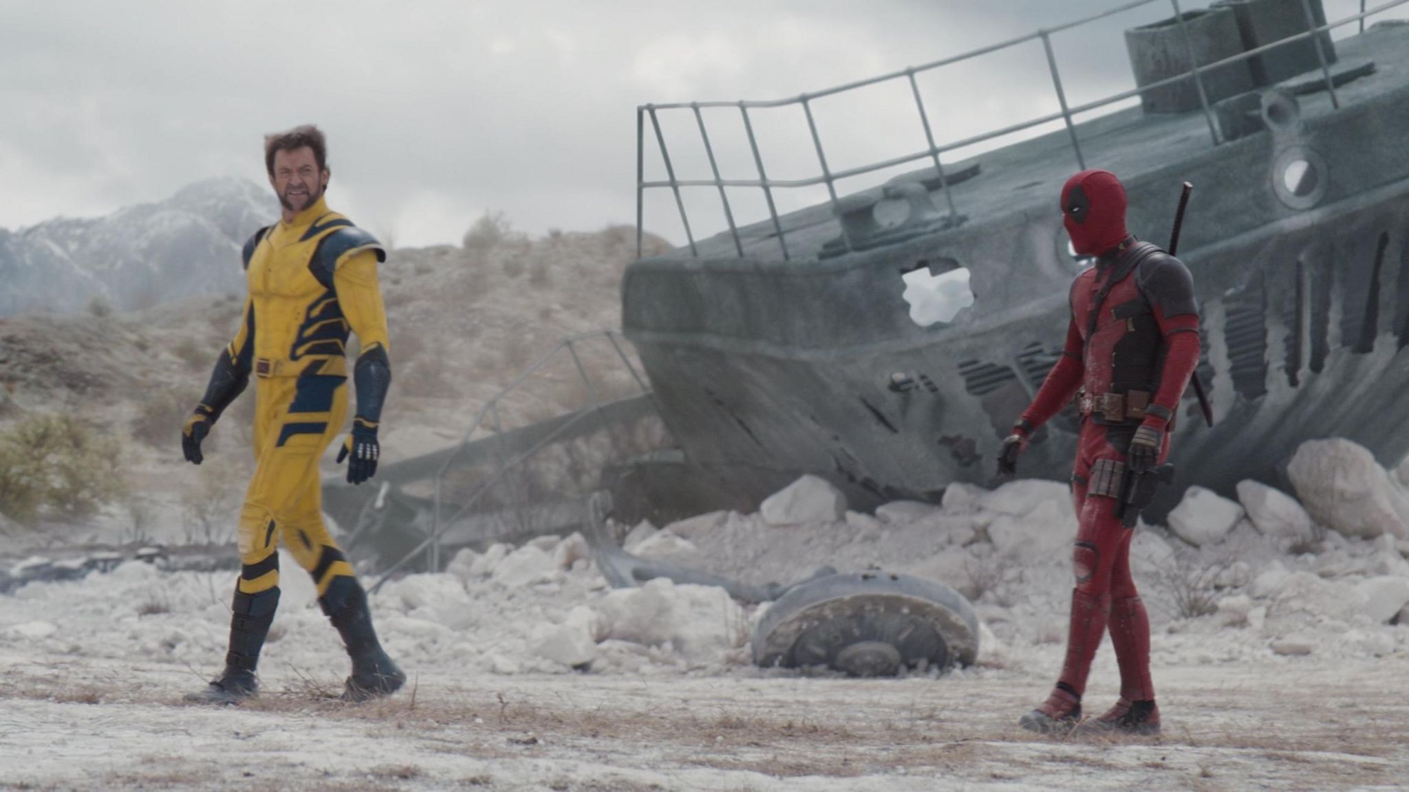 An image from the new deadpool and wolverine film. Jackman is on the left in his yellow wolverine outfir, whilst Ryan Reynolds is on the right in his red and black deadpool costume. In the background there is an old ship and it is some sort of dust plain.