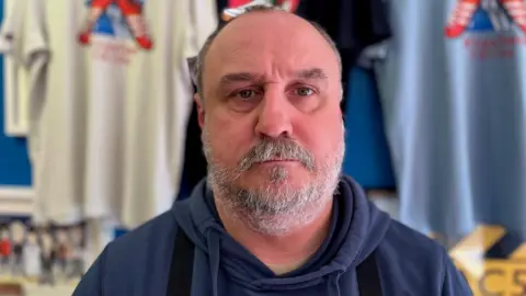 Steve Tapp standing in front of a rack of t-shirts at his shop, Wrexham Trainer Revival. He is middle-aged with a white beard.