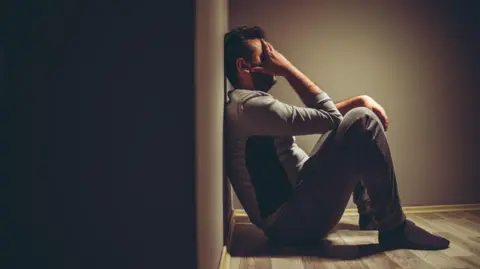 A stock image of man looking sad, sitting on the floor with his back against a wall