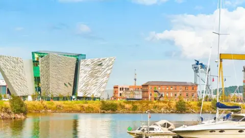 Getty Images Titanic Quarter in Belfast. Sea in the foreground. To the right, a partially visible view of a large, yellow crane, and left across the frame, a brick building, the Titanic Belfast building and industrial buildings.