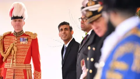 Leon Neal The UK Prime Minister smiles as he lines up alongside men in various uniforms.