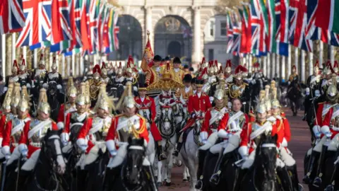 Getty Images A huge amount of horses are ridden by soldiers down the Mall in London. British flags line the street on both sides. The royal carriage is in the middle.