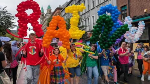 Group of people in colourful dress taking part in a parade carrying balloons in rainbow colours that spell out 'Rainbow'