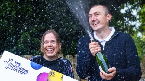 PA Katherine and Graeme White spraying fizzy wine from a bottle with a National Lottery sign