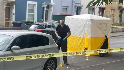 A police officer at the scene with yellow cones on the floor. He is standing with blue gloves and a face mask on. A yellow and white police tent is pictured behind him and there is a police cordon around the street.