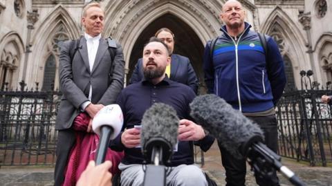 Martin Hibbert, sitting in a wheelchair, speaks to reporters outside the Royal Courts of Justice, with a number of microphones in front of him and three people behind him