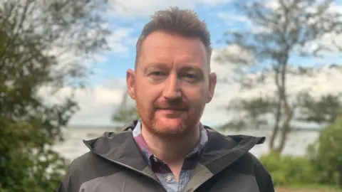 A man is looking directly into the camera. He has red hair and a beard. He is wearing a grey coloured waterproof jacket and a purple and blue checked shirt. Behind him you can see green trees and water.