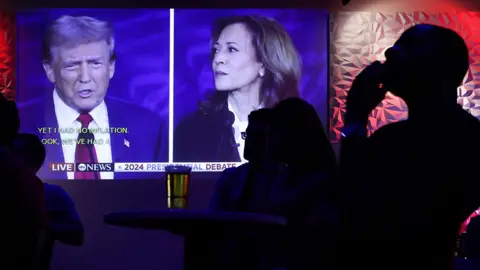 Donald Trump and Kamala Harris speak to each other during a debate televised on a screen at an event with the silhouette of a person in the foreground