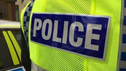 BBC Close up of an officer wearing a high vis vest with 'police' written on the back