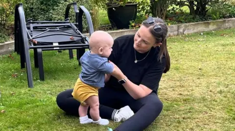 Stacey sitting on the grass in a garden. She is holding Frankie's hands while he is standing up. He is about four months old and wearing a yellow baby grow with a blue top over it. 