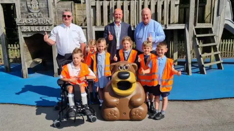 Six children in orange hi-vis vests, three men and a big plastic bear in a playground, all holding up their thumbs