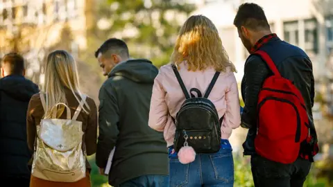 Stock photo showing students gathering and conversing outside a university building on a sunny day