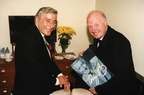 Tony Bennett and John Bennett sat on chairs with yellow flowers on a side table behind them, John holding a copy of Tony's record, both men in black suits smiling at camera