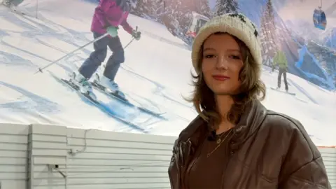 A teenager wearing a brown jacket and a white wooly hat smiles at the camera. Behind her is a large poster of people skiing