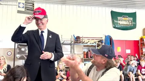 Joe Biden puts on a Trump 2024 hat surrounded by people applauding him