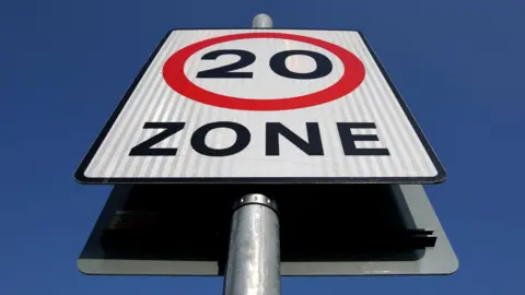 A white signboard with text saying 20 in black, surrounded by a red circle and the word Zone below it