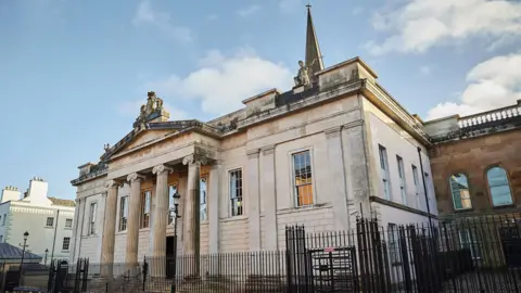 Crown court in Londonderry pictured with a black fence around the exterior. It is a stone building.