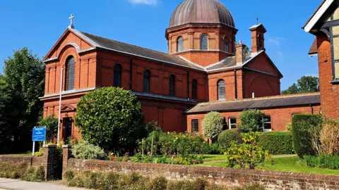 Fi Fi A sunny scene is dominated by a red brick church-type building. It has a dome at the rear and there is a cross at the top of the roof at the front. A garden runs along the building at the front.