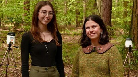 Two women in their twenties stand in front of two air sampling machines surrounded by woodland.