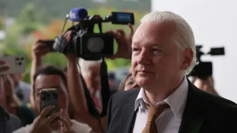 Julian Assange in a suit and tie surrounded by journalists and cameras