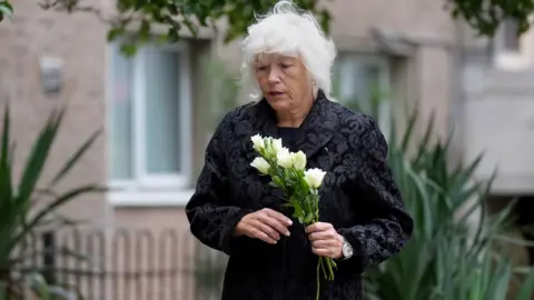 PA Melanie Leahy carrying white roses in her hand outside Chelmsford Civic Centre where the Lampard inquiry is being held. She is looking down and dressed in a black, long sleeved coat. Her hair is grey and shoulder length. She is about to place the flowers on a poster of her son placed on the ground alongisde photos of other people that have died under the care of mental health services in Essex