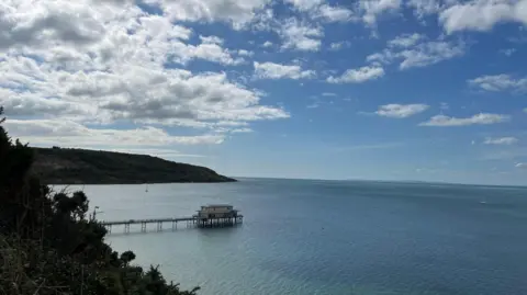 A sunny sky dominates this image of a structure standing out in a bay. Cliffs can be seen in the distance above the blue waters.