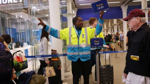 Male member of staff in train station wearing a yellow and blue high-vis jacket, holding his arms out and holding a sign saying Paris 12:01