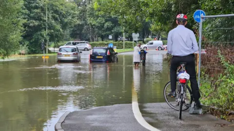 A flooded road with two cars stuck in the middle of it. A man on a bike is surveying the scene on a pavement above the water. Another woman is trying to push her bike through the flooded road