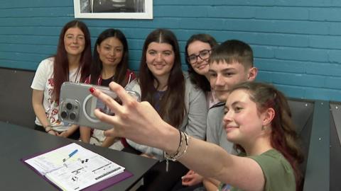 A group of six teenagers are sitting in a row. One is holding up a phone and taking a selfie of the whole group.