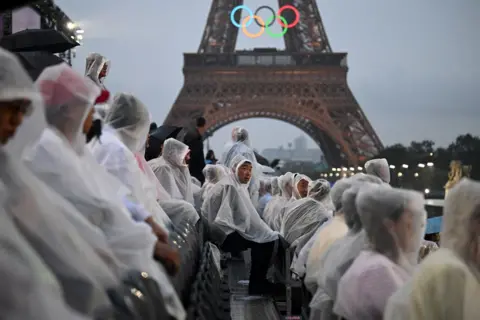 Oli Scarff/AFP Attendees wear rain covers as they sit in the stands in front of the Eiffel Tower